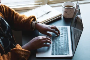 Image shows hands on a laptop with a coffee mug and notebook next to it.