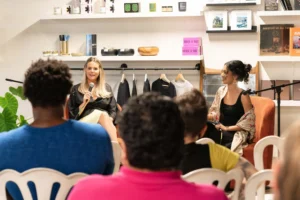 Image shows a group of people listening to two women with microphones present information. Everyone is seated.